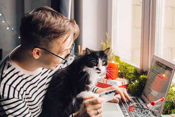 excited young man with black cat holding credit debt card shopping online purchase xmas presents on laptop home. Christmas gift box, New Year winter sale discount. Safe ordering delivery at quarantine