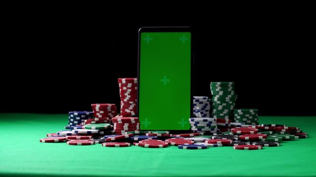 Green screen smartphone in vertical orientation in the middle of poker chips. Concept of playing at hazard games in online casino. Winning and losing money. Gambling as entertainment at leisure time.