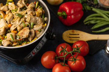The pan with frying meat and mix of vegetables on the stove near ingredients on dark blue background. Backstage of cooking pasta. Traditional italian cuisine. Food concept. Flat lay. View from above