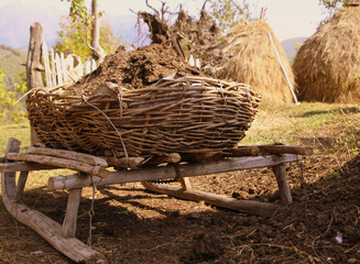 Summer time village life on The Caucasus Mountains. Authentic rural view of wicker basket full of dung stand on wooden sled and haystack background. 