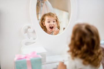 Reflection in the mirror of little screaming girl holding lip gloss brush in front of her
