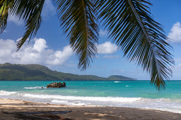 Panoramic view of the paradise beach with palm tree leaves, lone cliff (rock) in blue water of Atlantic Ocean and green mountains at the background, Rincon, Samana, Dominican Republic 