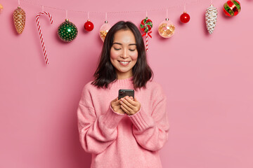 Brunette Asian woman with eastern appearance holds modern mobile phone for sending text messages connected to wireless internet views photos after New Year party poses over christmas decorations