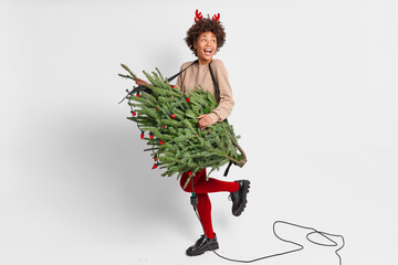 Winter and Christmas holidays concept. Positive dark skinned woman with curly hair wears bright red tights and shoes going to decorate New Year fir tree pretends to be rocker isolated on white wall