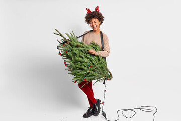 People entertainment and festive mood concept. Joyful Afro American woman pretends being rocker holds green fir tree with garlands as if guitar wears fashionable clothes and red reindeer horns