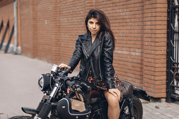 Naklejka na ściany i meble Portrait of a bright and daring adult model in a leather jacket and dress sitting on a black motorcycle and looking straight at the camera against the backdrop.