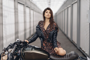 Fototapeta na wymiar Portrait of a lovely young lady in a leather jacket and dress posing next to a black motorcycle and looking straight into the camera against the backdrop of white walls. Sexuality concept