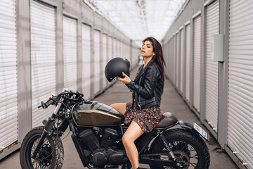 Fototapeta na wymiar Girl on a motorcycle. Young sexy girl in a leather jacket and dress sitting on her retro motorcycle and holding a helmet in her hands is getting ready to drive. Extreme ​​concept