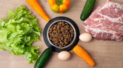 Dry pet dog food with natural ingredients. Raw meat, vegetables, eggs and salad near bowl with dry pet feed on wooden background. concept of pet correct balanced and healthy nutrition