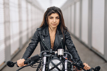 Fototapeta na wymiar Portrait of a cheeky bright dark-haired model in a black leather jacket sitting on a black retro motorcycle and looking straight into the camera against a white background. Beauty concept