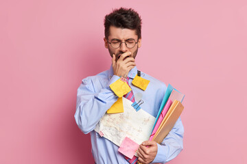 Tired student has sleepy expression covers mouth with hand and yawns dressed in formal clothes prepares for exams poses against pink background. Executive worker makes memo notes for project