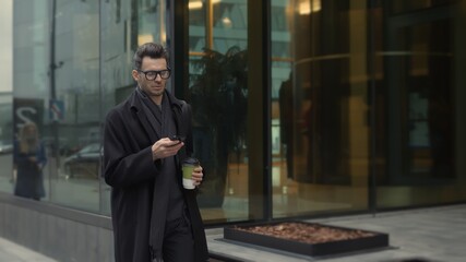 Man in black coat and eyeglasses looking at the phone walking along the street with coffee, slow motion. City street with glass windows of office building on background