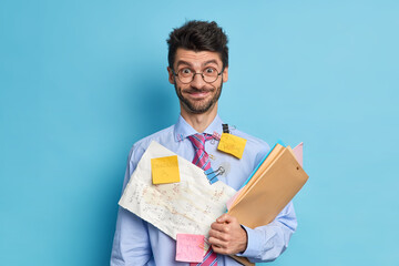 Cheerful bearded male financier prepares stratup project poses with papers and folders dressed in...