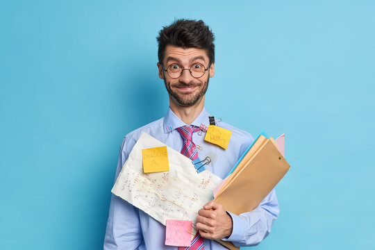 Cheerful bearded male financier prepares stratup project poses with papers and folders dressed in formal clothes wears round spectacles isolated over blue background. Office worker being busy