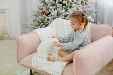 Obraz na płótnie Canvas A three-year-old girl in a dress sits in a pink armchair on a New Year tree. A child plays with a polar bear in a light interior. Christmas and New Year concept.