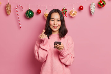Lovely Asian woman shapes mini heart with fingers expresses love holds mobile phone for surfing in social networks makes shopping online and buys Christmas presents for family poses at home. - Powered by Adobe