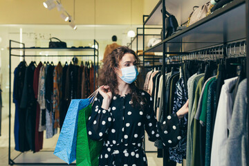 Young beautiful woman in a protective mask on her face, chooses clothes in the store, makes safe...