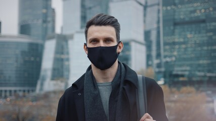 Fototapeta na wymiar Man in black coat and black protective mask on background of business skyscrapers, looking at the camera. Gimbal shot of businessman in coat standing near international business centre