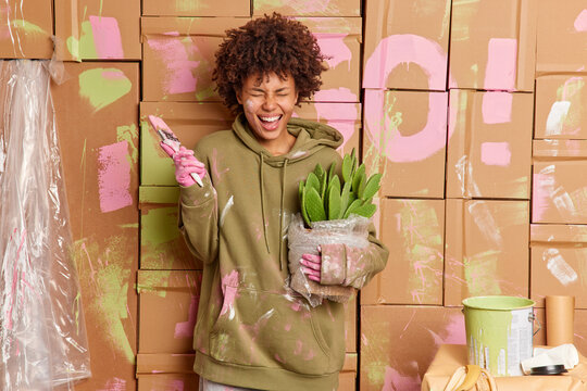 Overjoyed female house owner has fun while refurbishing walls of house holds pot of cactus paint brush laughs happily finishes room renovation dressed in casual sweatshirt. Home repair concept