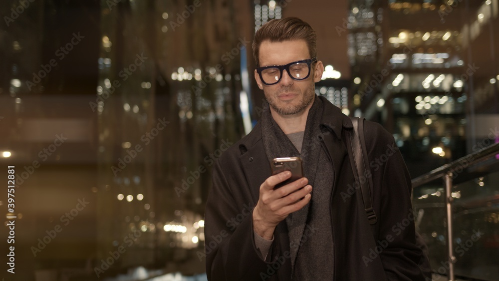 Poster man in eyeglasses with phone in hand on background of building with lights on. handheld night shot o