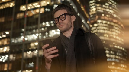 Man in eyeglasses with phone in hand on background of building with lights on. Handheld night shot of caucasian man texting in phone near business building with cars