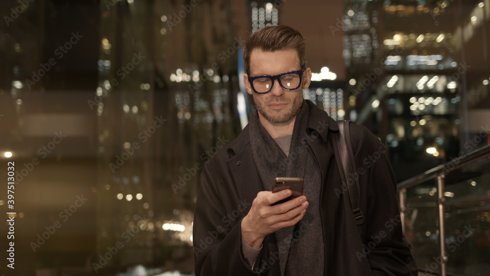 Poster man in eyeglasses with phone in hand on background of building with lights on. handheld night shot o