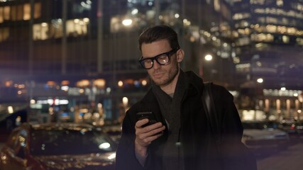 Fototapeta premium Man in eyeglasses with phone in hand on background of building with lights on. Gimbal night shot of caucasian man texting in phone near business building