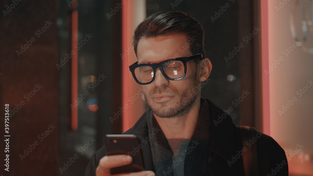 Fototapete man in eyeglasses with phone in hand on background of building with lights on. handheld night shot o