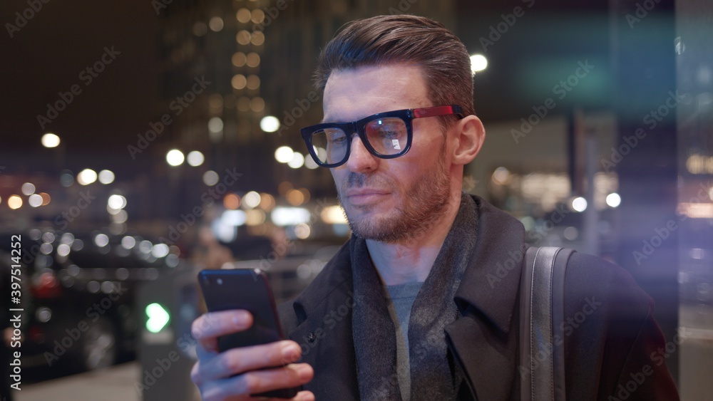 Fototapete Man in eyeglasses with phone in hand on background of building with lights on. Gimbal night shot of caucasian man texting in phone near business building with cars