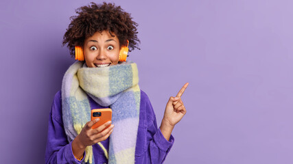 Happy dark skinned woman has curly bushy hair wrapped in warm winter scarf holds mobile phone for online communication wears headphones on ears surprised to see amazing offer points on right