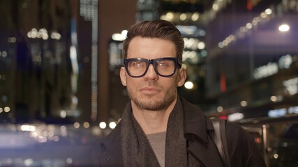 Obraz premium Man in eyeglasses standing on background of business skyscrapers, windows with lights. Portrait gimbal shot of businessman in coat standing near business centre downtown at night