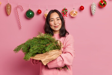 Charming female with dark hair and pleasant smile embraces fir tree branches arranged in bouquet...