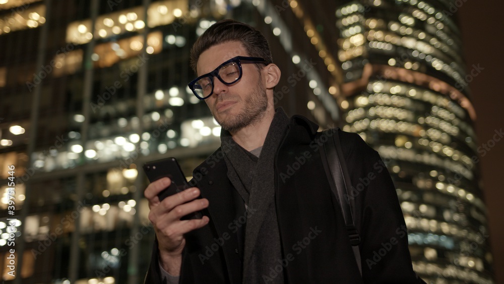 Poster man in eyeglasses with phone in hand on background of building with lights on. handheld night shot o