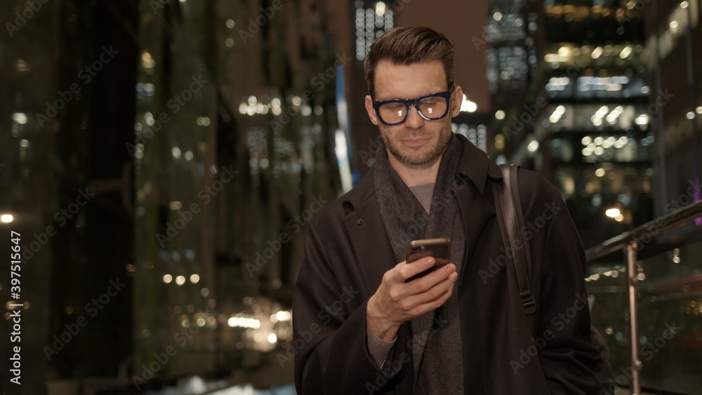 Poster man in eyeglasses with phone in hand on background of building with lights on. handheld night shot o