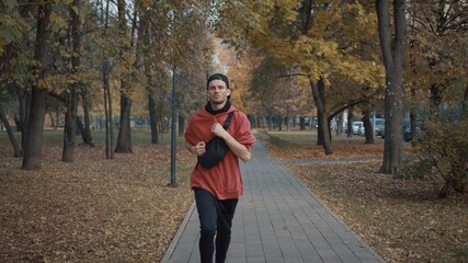 Man running in the park slow motion, young man in sport clothes and cap listening to music running through the park in the autumn day on a track between trees