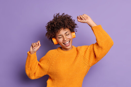 Positive dark skinned young woman dances carefree wears orange jumper moves with rhythm of music uses stereo headphones isolated over purple background. People hobby and entertainment concept
