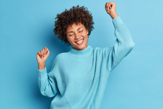 Joyful carefree woman poses full of happiness raises arms and dances with satisfaction smiles pleasantly and closes eyes dressed in long sleeved jumper isolated over blue background has upbeat mood