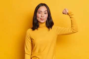 Fotobehang Serious self confident brunette young Asian woman raises hand up and shows demonstrates strength looks like strong powerful bodybuilder dressed in casual turtleneck isolated over yellow background © Wayhome Studio