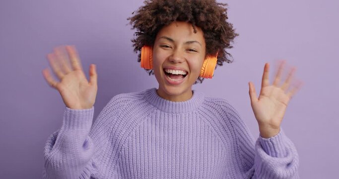 Overjoyed happy woman waves hello gesture glad to meet best friend welcomes someone with hospitable expression wears wireless headphones on ears dressed in casual sweater poses indoor over purple wall