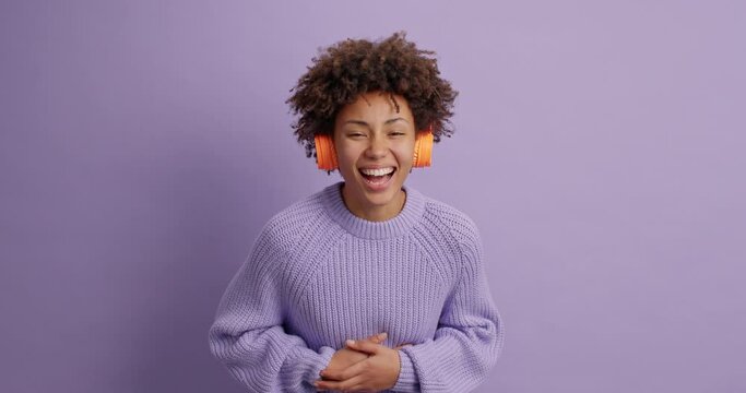 Sincere dark skinned woman cannot stop laughing holds stomach hears funny anecdote smiles broadly dressed in knitted sweater listens audio track via headphones isolated over purple background