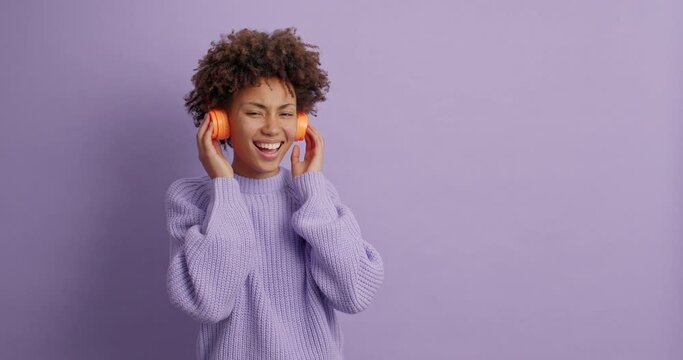 Optimistic curly woman moves carefree against purple background enjoys favorite melody catches every bit of song has good mood poses in studio with blank space on violet wall for your promotion