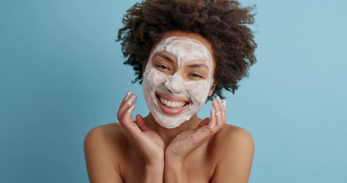 Happy young African American woman with curly hair looks gladfully at camera applies nourishing beauty mask on face cares about skin smiles broadly has naked body poses against blue background