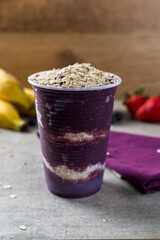 Brazilian frozen açai berry ice cream on a plastic cup with oatmeal flakes. bananas, strowberries and blueberries on background. wood background. Summer menu front view