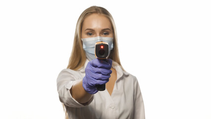 Nurse in protective mask pointing sensor thermometer on camera wearing white medical hospital robe. Young blond haired nurse checking on patients or coworker temperature.