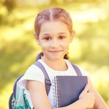 Young pretty girl holding books. School education concept. High quality photo. Little student with backpack. Lifestyle kindergarten American kid. Green outdoor background. Summer or fall park