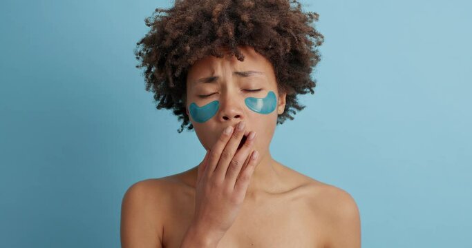 Dissatisfied sad woman has curly hair yawns and covers mouth with hand stands shirtless indoor applies collagen patches under eyes has bare shoulders isolated over blue background. Eye mask.