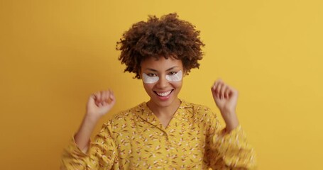 Young carefree woman enjoys beauty treatments wears collagen patches under eyes dances positively against yellow background dressed in casual night costume. Anti aging and skin care concept.