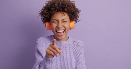 Cheerful dark skinned woman appears suddenly on purple background raises palms says boo and tries to frigten someone then smiles and has fun with friend wears headphones on ears casual jumper