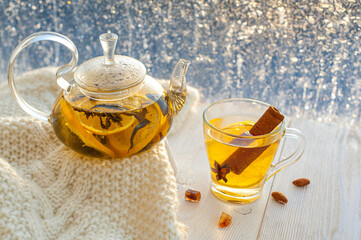 Winter warming tea with lemon and orange against the background of frosty patterns. Hot drink in a glass kettle and a cup on a wooden table in the background of the window.