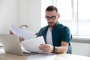 Focused young Caucasian male employee sit at desk at home office work on computer reading paperwork. Concentrated man worker busy using laptop consider financial documents at workplace.